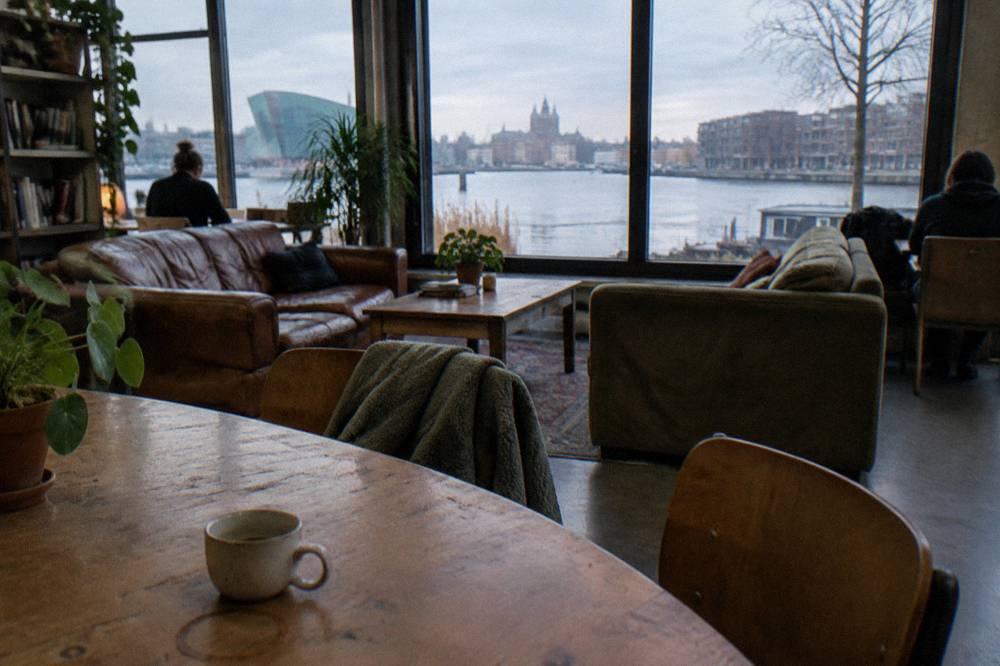 Modern hostel common room with wooden tables, sofas and large windows overlooking water