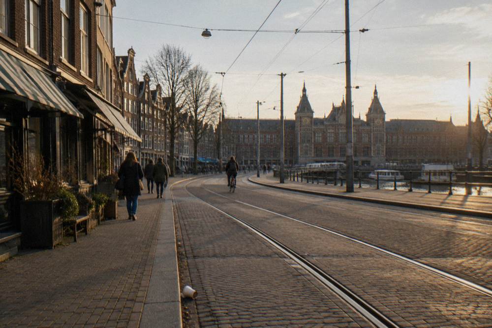 An Amsterdam street corner with a tram line, brick facades and shop fronts in late afternoon light