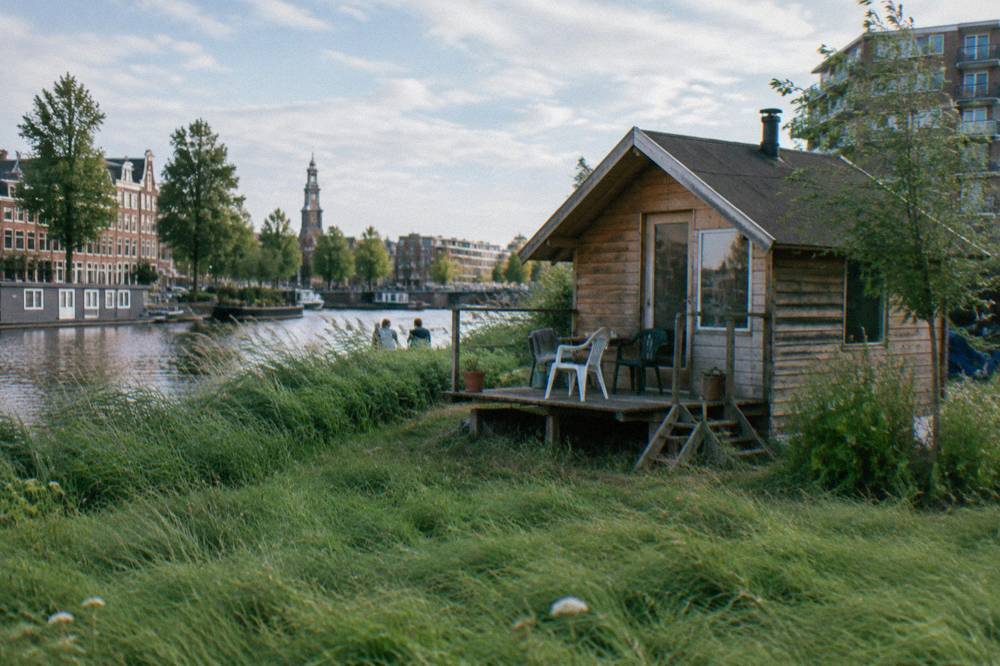 Small wooden cabin with a pitched roof beside a canal and open grass at a city campsite