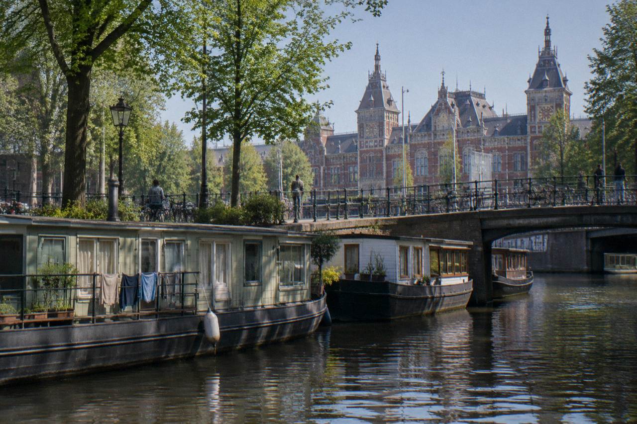 A canal in Amsterdam lined with moored houseboats beside a tree-shaded footbridge